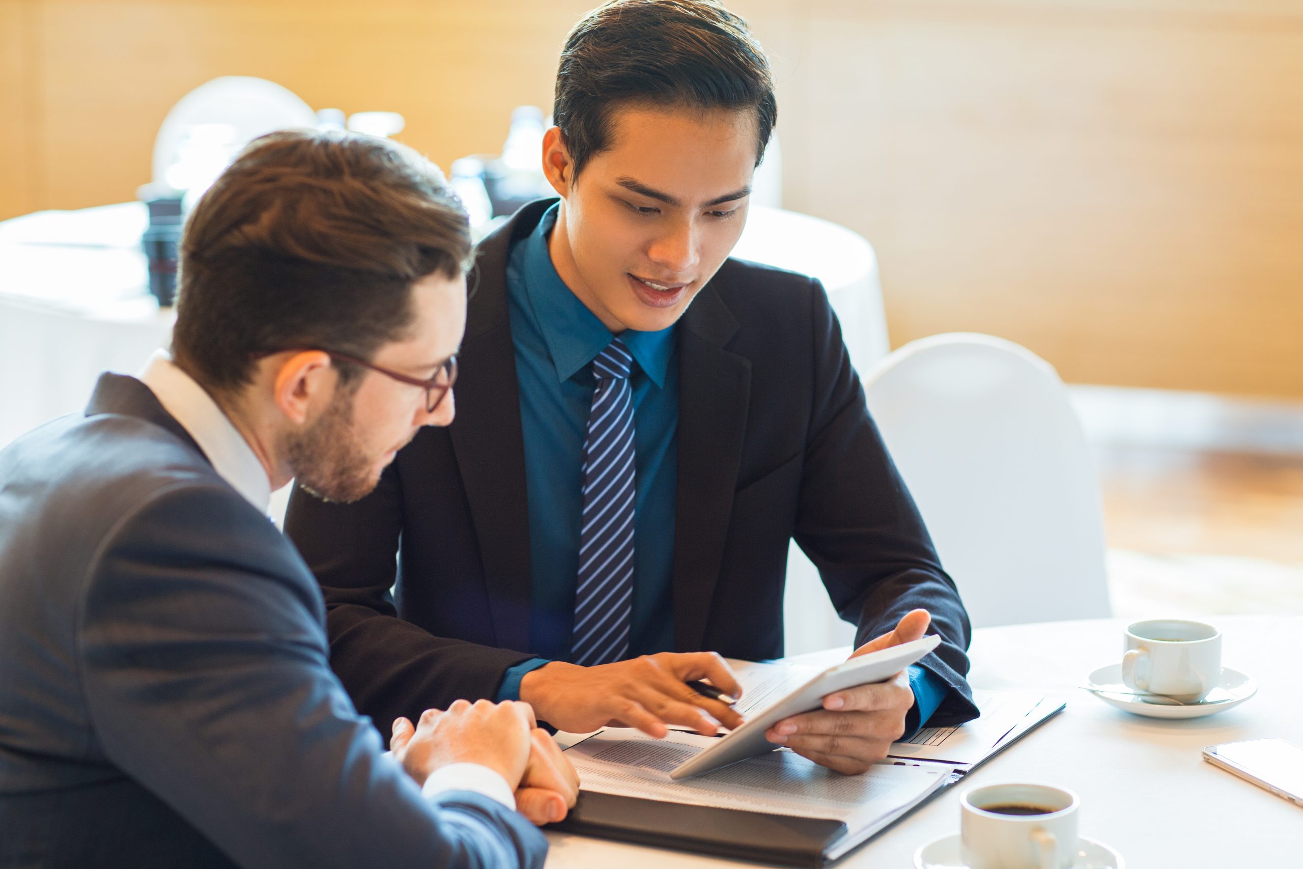 Closeup of two content adult business men sitting at table, using tablet computer, working with documents and discussing them in cafe